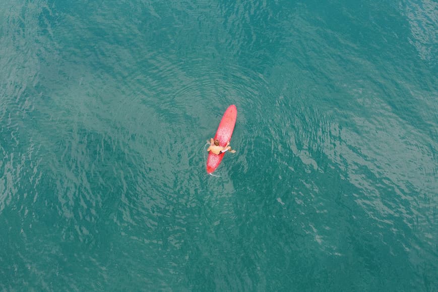 Aerial view of a surfer paddling on a red board in turquoise water