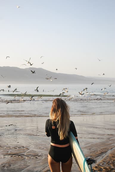 Surfer walking toward the waves in Imsouane Morocco with seagulls flying over the beach