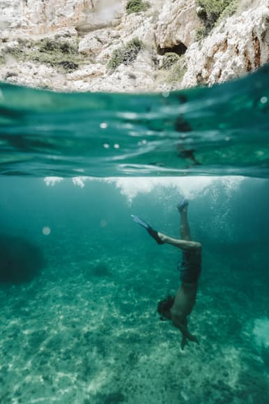 Person diving underwater in clear turquoise water near rocky Mediterranean cliffs